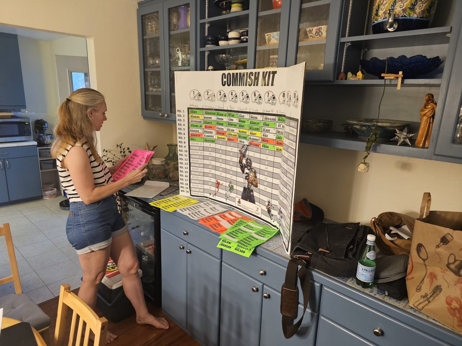 A woman standing at the counter reviewing colored player sticker sheets next to the partially filled draft board